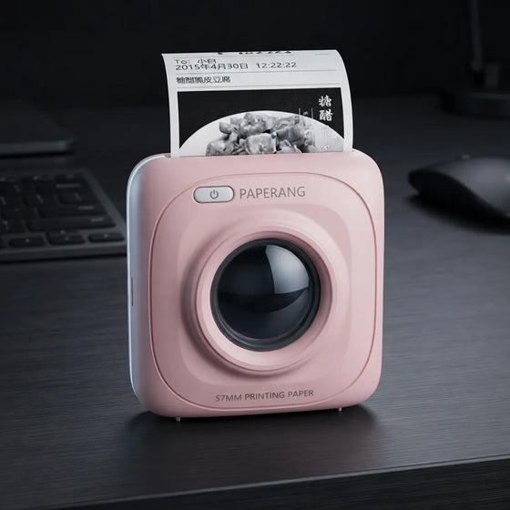 A pink SQUARE paperless printer sits on a dark desk with a printed sheet emerging from the top. The device has a glossy black circular lens area, a power button, and the brand name PAPERANG printed above it.