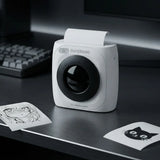A compact white Paperang portable printer with a black circular lens area sits on a dark desk, with printed sheets and a blurred keyboard in the background.