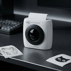 A compact white Paperang portable printer with a black circular lens area sits on a dark desk, with printed sheets and a blurred keyboard in the background.