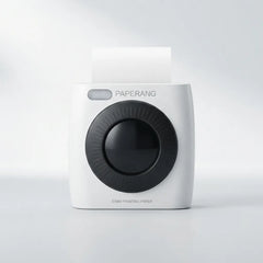A Paperang instant camera-style printer in white with a large black circular lens area and a sheet of white paper emerging from the top, shown on a clean light gray studio background.