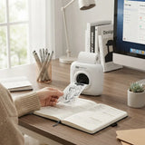 A white HP Pango instant photo printer on a wooden desk, with a hand holding a printed sheet coming out of the slot. The scene includes a computer monitor, books, a lamp, a cup of pens, and a small succulent.