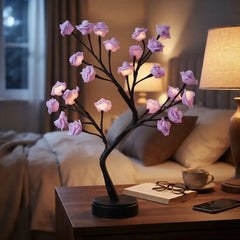A decorative tabletop lamp shaped like a flowering branch with glowing pink rose-shaped bulbs, placed on a wooden nightstand in a softly lit bedroom setting.