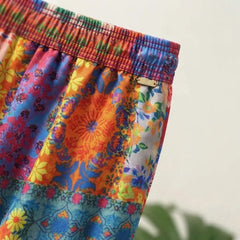 A close-up of a colorful floral print garment, likely a skirt or shorts, featuring a vibrant patchwork design with orange, blue, pink, yellow, and green patterns. The waistband is gathered with a matching patterned elastic.