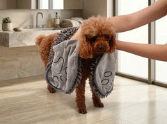 A small brown poodle stands on a bathroom floor while a person holds it and drapes a gray microfiber pet wash blanket with black paw-print outlines over its body. A marble sink and bright windows are visible in the background.