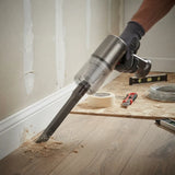 A cordless handheld vacuum cleaner with a metal floor nozzle being used to clean wood flooring in a room with an unfinished wall and baseboard. A gloved hand holds the vacuum, with construction tools like tape, a utility knife, and wire visible on a plywood sheet in the background.