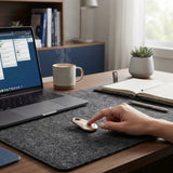 A person's hand rests on a small beige personal alarm lying on a dark gray felt desk mat on a wooden desk, with a laptop, mug, notebook, and plant in a home office setting.