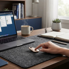 A person's hand rests on a small beige personal alarm lying on a dark gray felt desk mat on a wooden desk, with a laptop, mug, notebook, and plant in a home office setting.