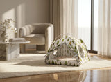 A small dome-style pet bed with a green and white tree pattern sits on a cream rug in a bright, modern living room with a chair, side table, and large window.