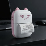 A small portable desktop printer with a cute cat-face design, featuring pink ears and a pink side panel on a white body. A sheet of paper is emerging from the printer, which is placed on a dark desk next to a laptop and mouse.