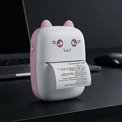 A small portable desktop printer with a cute cat-face design, featuring pink ears and a pink side panel on a white body. A sheet of paper is emerging from the printer, which is placed on a dark desk next to a laptop and mouse.