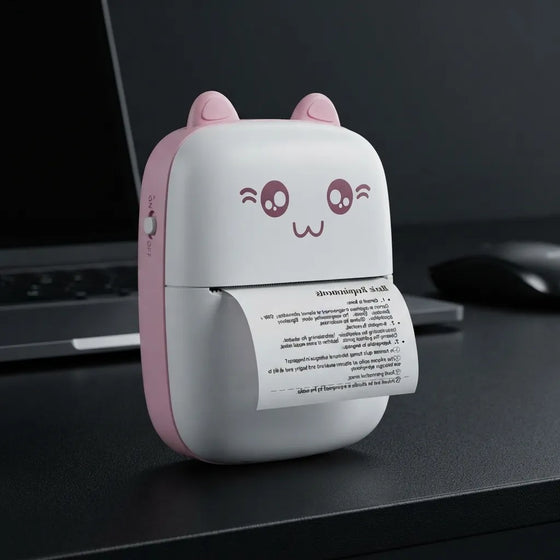 A small portable desktop printer with a cute cat-face design, featuring pink ears and a pink side panel on a white body. A sheet of paper is emerging from the printer, which is placed on a dark desk next to a laptop and mouse.