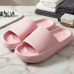 A pair of light pink textured slide slippers on a white marble surface, with folded towels and a soap dish in the background.