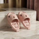 A pair of light pink shark-shaped slide sandals with cute cartoon eyes and teeth details, displayed on a white marble surface with a warm-toned background.