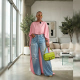 A woman stands in a modern living room wearing a pink draped top, light-wash distressed wide-leg jeans, and carrying a bright green handbag.