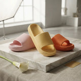 Three ribbed slide sandals in pastel pink, mustard yellow, and burnt orange arranged on a white marble slab in a bright, softly lit interior.