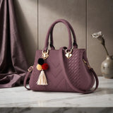 A structured handbag in dusty mauve purple with a quilted chevron pattern, dual top handles, a shoulder strap, and a decorative pom-pom tassel charm, displayed on a marble surface with a draped fabric backdrop.