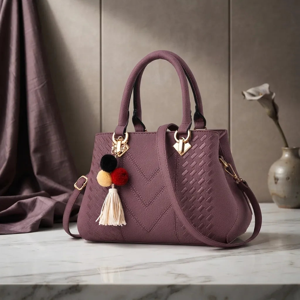 A structured handbag in dusty mauve purple with a quilted chevron pattern, dual top handles, a shoulder strap, and a decorative pom-pom tassel charm, displayed on a marble surface with a draped fabric backdrop.