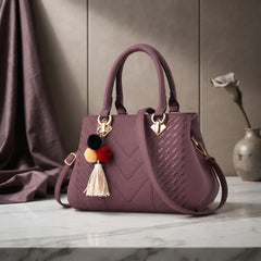 A structured handbag in dusty mauve purple with a quilted chevron pattern, dual top handles, a shoulder strap, and a decorative pom-pom tassel charm, displayed on a marble surface with a draped fabric backdrop.