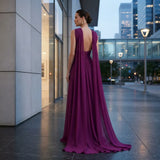 A woman wearing a floor-length magenta evening gown with a draped open back, photographed from behind in an urban plaza at dusk.