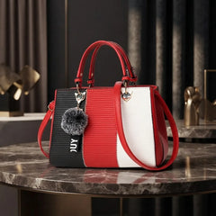 A structured handbag with a tri-color design of black, red, and white, featuring a red top handle, a gray pom-pom charm, and the brand name 'JUY' printed on the black side panel, displayed on a marble table.