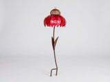 A metal flower garden stake sculpture with a vibrant red petal-shaped top, a brown mesh center, and a long brown stem with leaves, standing on a white studio background.