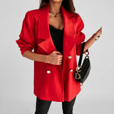 A woman wearing a bright red double-breasted blazer with gold buttons over a black top and black pants, holding a black handbag with gold hardware against a light gray studio background.