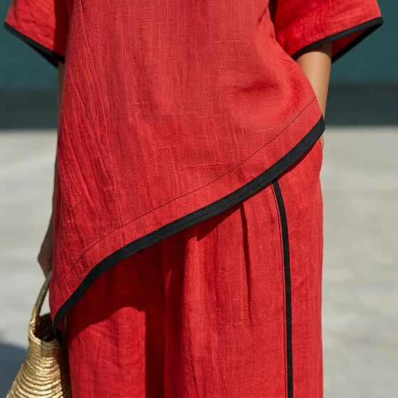 A close-up of a red linen-like dress or tunic with black trim along the asymmetrical hem and sleeve edges, worn by a model in an outdoor setting.