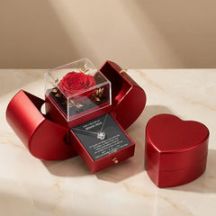 A red heart-shaped gift box set displayed on a light marble surface against a beige background. One box is open to reveal a preserved red rose in a clear cube and a necklace with a heart pendant on a card.