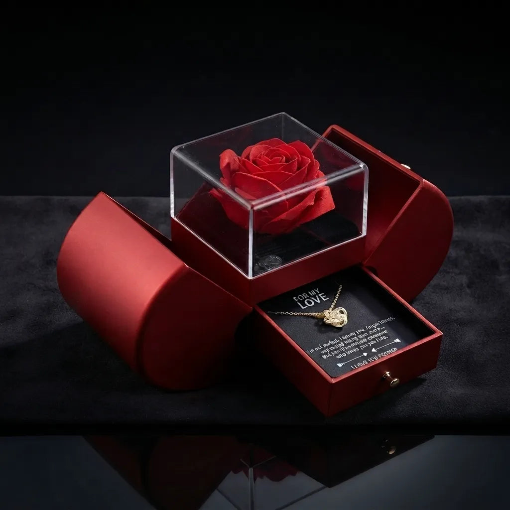 A red rose preserved in a clear acrylic display box, set inside an open red jewelry gift box with a necklace and message card inside, on a dark reflective surface.