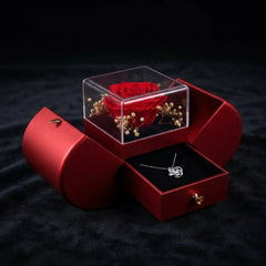 A red heart-shaped jewelry gift box on a black textured background, with the top clear cube lid open to show a preserved red rose and dried flowers, and the bottom drawer pulled open to reveal a silver necklace on a black insert.