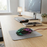 A gaming mouse with RGB lighting resting on a gray desk mat on a wooden desk, with a keyboard, notebook, and monitor in the background.