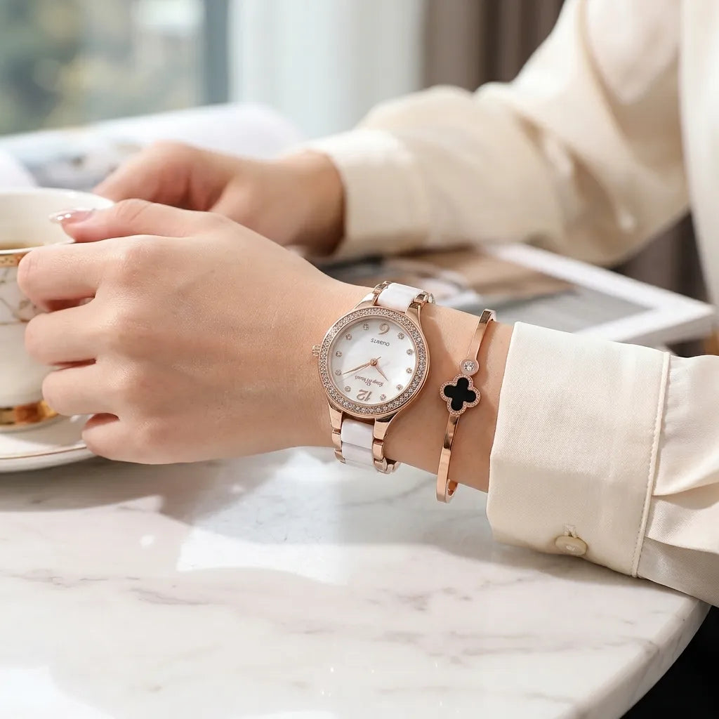 A woman's wrist wearing a white leather watch with a rhinestone-embellished rose gold case and a matching white leather bracelet, paired with a rose gold bangle bracelet featuring a black clover charm. She is holding a teacup on a white marble table.