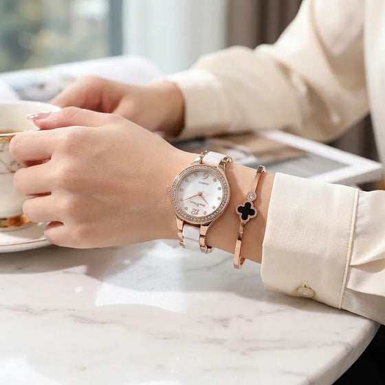 A woman's wrist wearing a white leather watch with a rhinestone-embellished rose gold case and a matching white leather bracelet, paired with a rose gold bangle bracelet featuring a black clover charm. She is holding a teacup on a white marble table.
