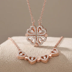 Rose gold jewelry set featuring a four-leaf clover pendant necklace and a matching heart-link bracelet, both adorned with clear rhinestones, displayed on a textured beige surface.