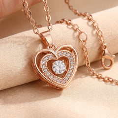 Rose gold heart-shaped pendant necklace with a clear central gemstone and small clear accent stones, displayed on a beige fabric surface.
