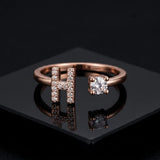 A rose gold open ring featuring a large pavé-set letter H and a single round-cut clear gemstone on the band, displayed on a glossy black surface.