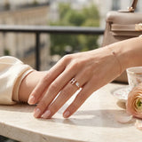 A close-up lifestyle shot of a person's hand resting on a marble table, wearing a delicate rose gold ring with an 'H' initial and a small clear gemstone. The background features a soft-focus view of a balcony railing, a beige handbag, and a pink rose on a table setting.