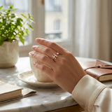 A close-up lifestyle shot of a person's hand wearing a delicate silver ring with a small heart and letter 'H' design, resting on a marble table next to a cup and notebook.