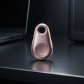 A small teardrop-shaped metallic tracker or key finder in a rose gold finish, standing upright on a dark reflective desk with a blurred laptop and computer tower in the background.