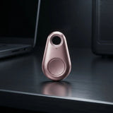 A small teardrop-shaped metallic tracker or key finder in a rose gold finish, standing upright on a dark reflective desk with a blurred laptop and computer tower in the background.