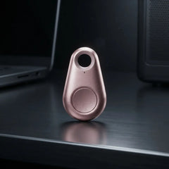 A small teardrop-shaped metallic tracker or key finder in a rose gold finish, standing upright on a dark reflective desk with a blurred laptop and computer tower in the background.
