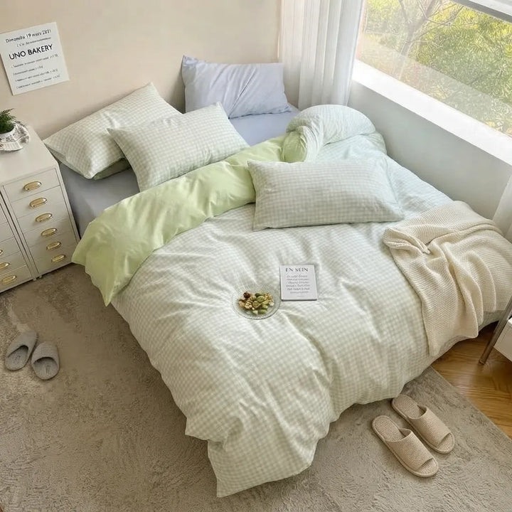 A neatly made bed in a bright bedroom with a light pastel bedding set. The duvet and pillows are in soft off-white and pale green tones with a subtle grid pattern, complemented by a cream-colored textured throw blanket.