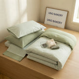 A neatly made bed with pale sage green and white gingham bedding, including pillows, a folded duvet, and a matching sheet, placed on a light wood bed frame in a softly lit room. A framed sign reading 'UNO BAKERY' stands behind the bed, and a small folded towel or cloth rests on top.