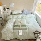 A neatly made bed in a bright bedroom setting, styled with a pale sage green and white gingham check bedding set. A folded matching pillowcase or sheet bundle tied with a cream ribbon sits on the bed, alongside a hanging product tag.