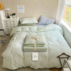 A neatly made bed in a bright bedroom setting, styled with a pale sage green and white gingham check bedding set. A folded matching pillowcase or sheet bundle tied with a cream ribbon sits on the bed, alongside a hanging product tag.