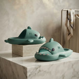 A pair of shark-shaped slide sandals in a muted teal-green color, displayed on a white marble pedestal with a textured beige background.