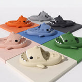 Six cartoon shark-shaped slide sandals arranged on a white background, each placed on a different colored square tile. The sandals are orange, gray, black, pink, blue, and green.