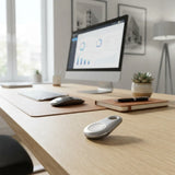 A silver oval smart key fob lies on a light wood desk in a bright home office setting, with a computer monitor, keyboard, mouse, notebook, and small succulent in the background.
