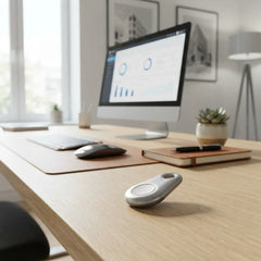 A silver oval smart key fob lies on a light wood desk in a bright home office setting, with a computer monitor, keyboard, mouse, notebook, and small succulent in the background.