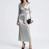 A woman models a long-sleeve wrap midi dress in a light silver-gray satin fabric, styled with a black clutch and black heels against a plain light gray studio background.